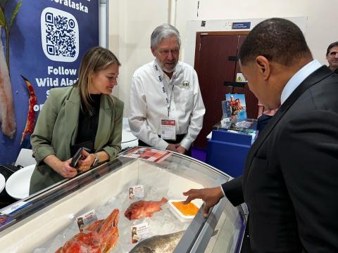 Administrator B. Daniel Whitley, dressed in a suit, stands to the right of exhibitors from Alaska Seafood at Gulfood 2025. The group is smiling, and behind them, a booth showcases images of fresh seafood and branding for U.S. seafood exports